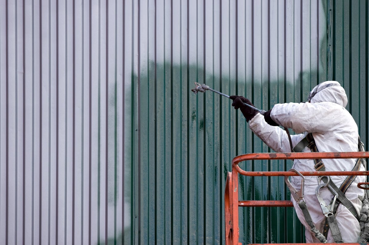 Person in protective gear spray painting a corrugated metal wall green from an elevated platform