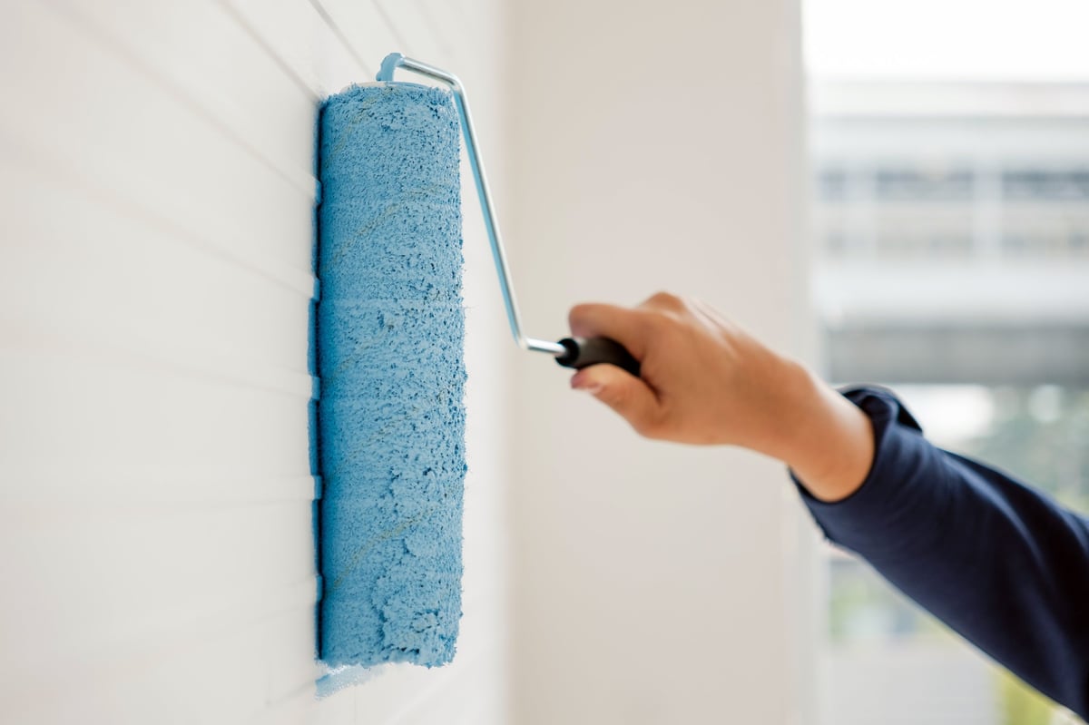 Hand holding a paint roller applying bright blue paint on a white textured wall