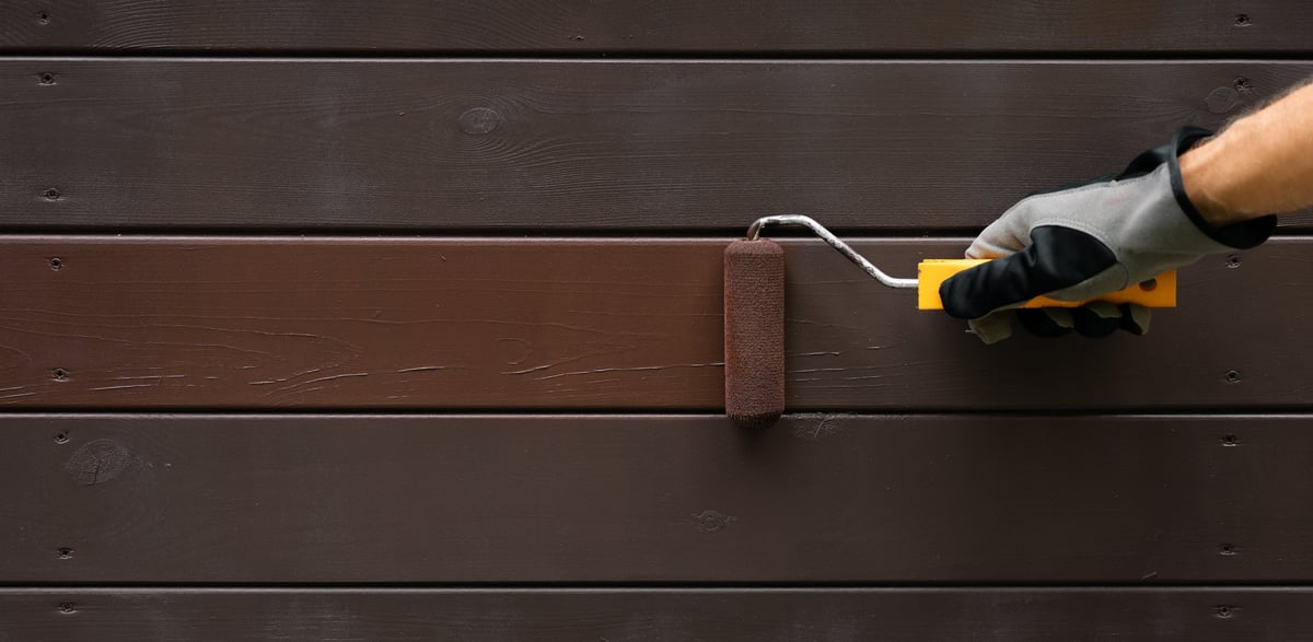 hand with paint roller painting wooden house exterior siding with protective color