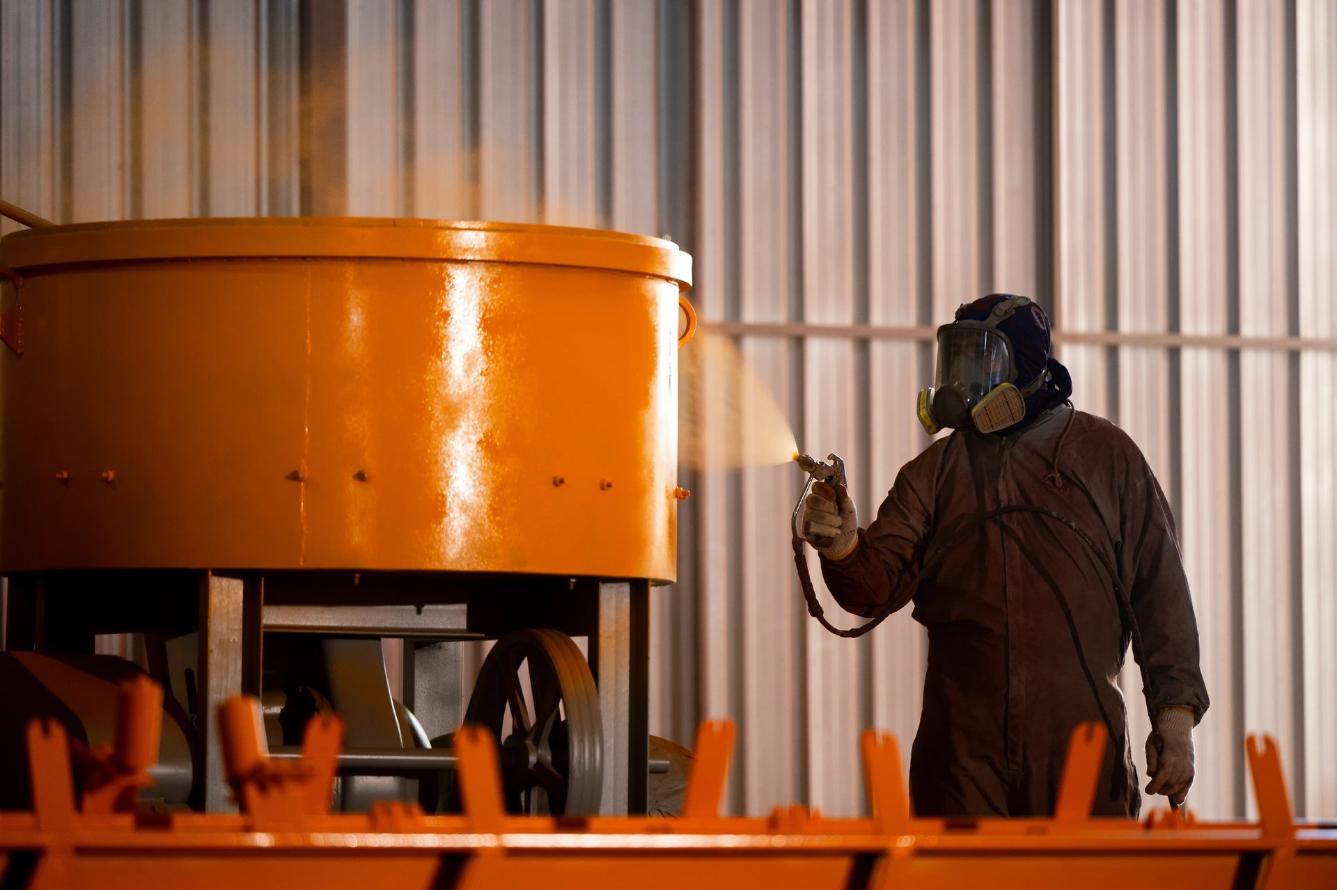 Industrial painter spray painting yellow coating on heavy machinery metal parts wearing protective suit and respirator