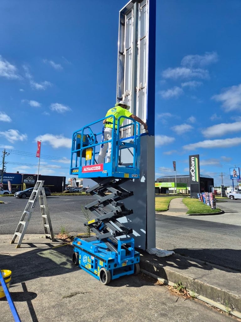 Blue and black scissor lift with elevated platform and worker in high-visibility gear on a parking lot under clear blue sky