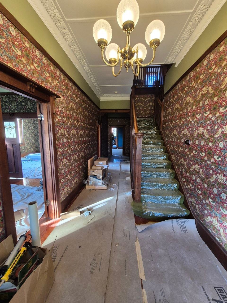 Historic hallway with ornate floral wallpaper, brass chandelier, wooden railings, and staircase under renovation