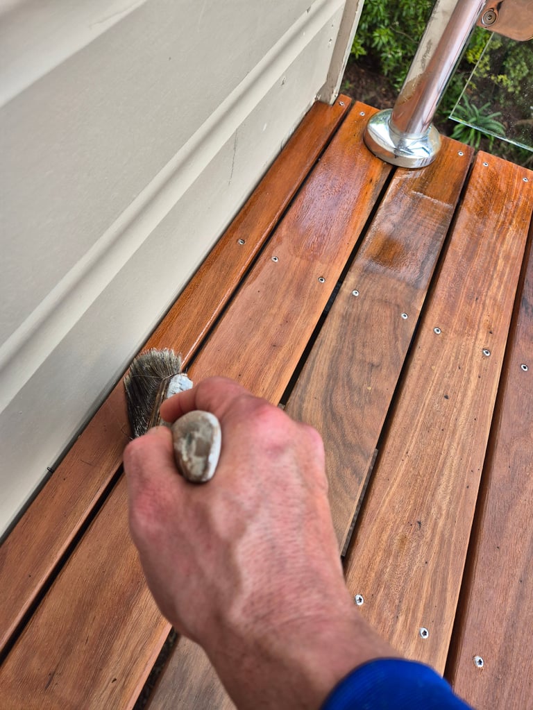 Person holding a brush over wooden deck boards next to a white railing and metal post
