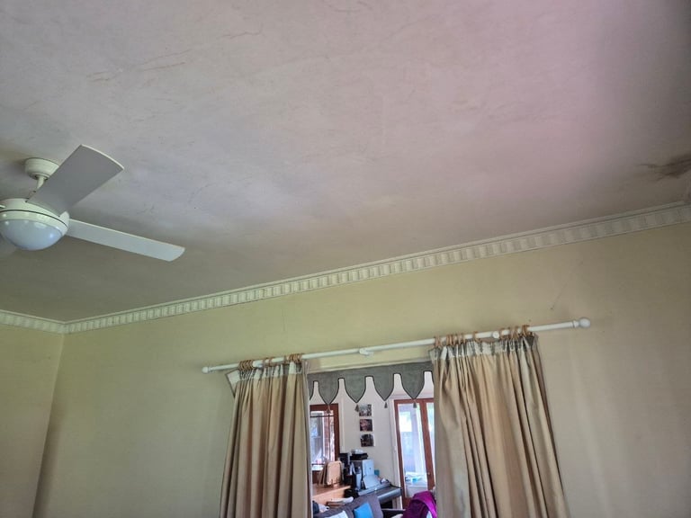 Interior bedroom with beige walls, ceiling fan, and beige curtains hung on a metal rod covering a window