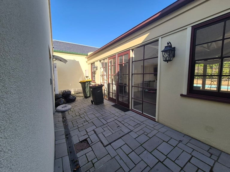 Sunny residential patio with paved flooring, cream-colored walls, black-framed glass doors and windows, and a wall lamp