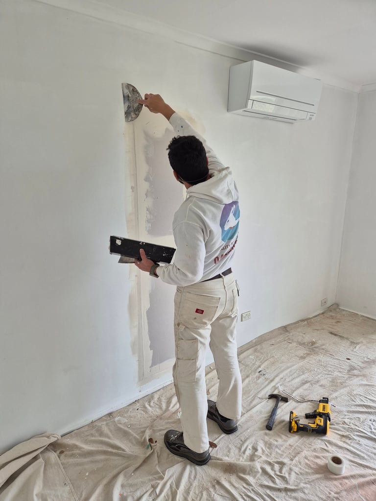 Worker in white protective gear and mask using a putty knife on a wall during interior renovation, with drop cloths and tools visible.