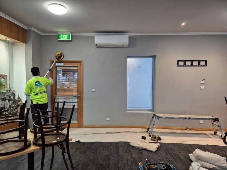Worker in yellow safety vest installing wall-mounted air conditioning unit in an empty indoor space with furniture and tools scattered on floor