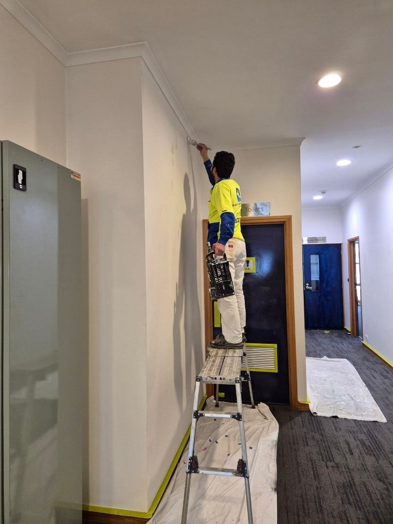 Painter in yellow shirt on ladder painting a white interior wall