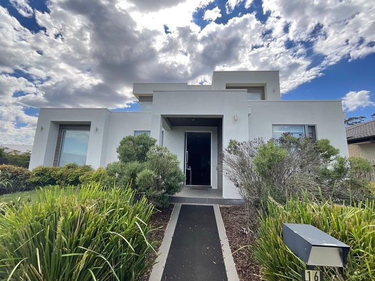 Modern white minimalist house with flat roof, black entrance door, and landscaped front garden under cloudy blue sky