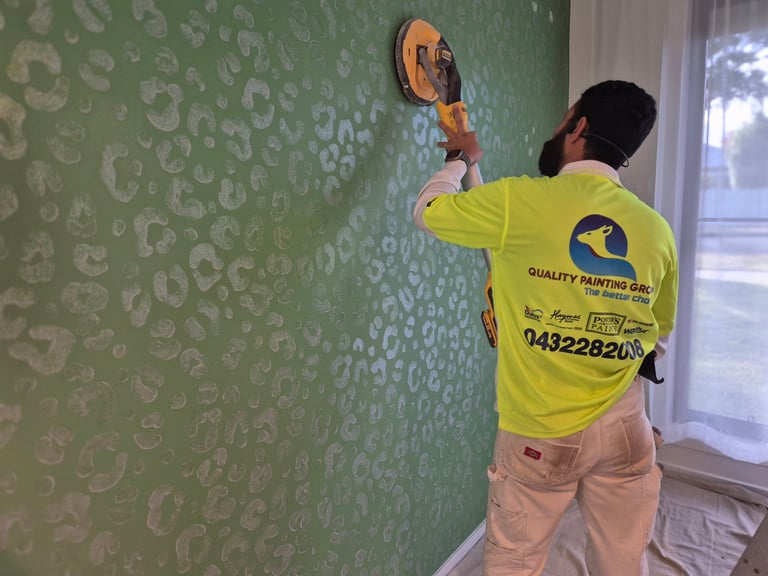 Worker in yellow shirt removing wallpaper from green patterned wall with circular scraper tool