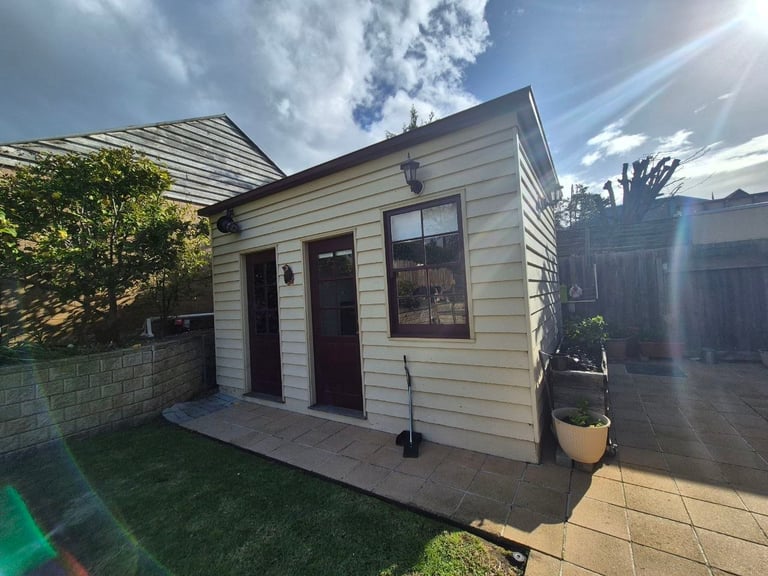 Small modern backyard shed with cream siding, dark door and window, concrete porch, and manicured lawn on sunny day