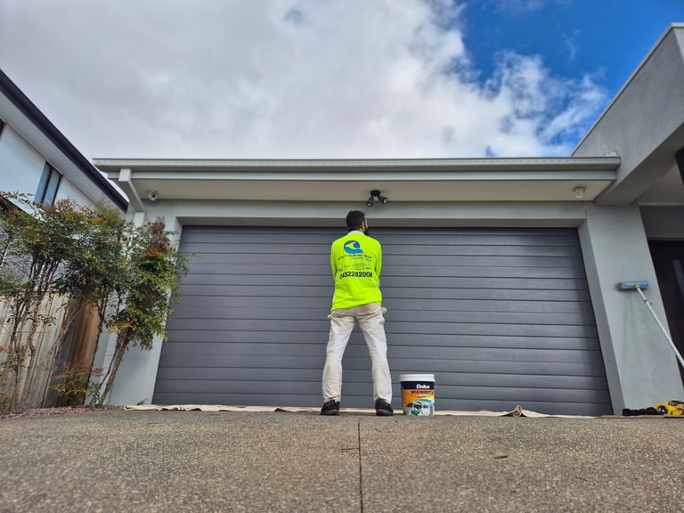 Worker in yellow safety vest painting gray garage door on modern residential home