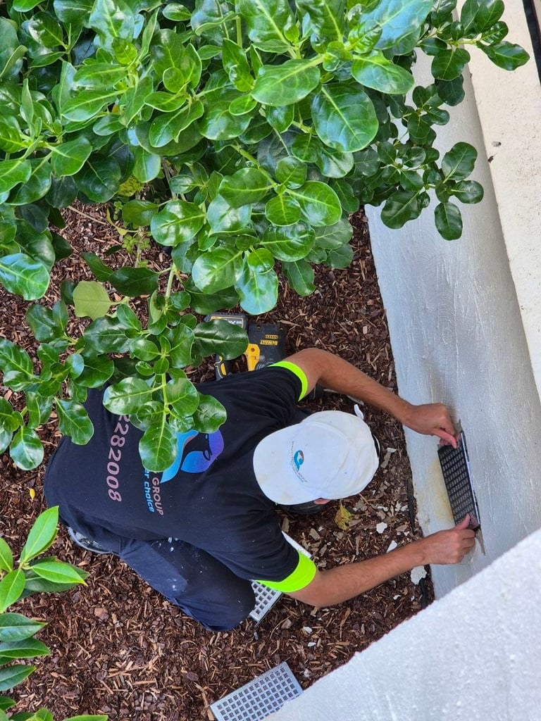 Person in black and yellow athletic wear kneeling by potted green plants while holding a white water container in a mulched garden bed