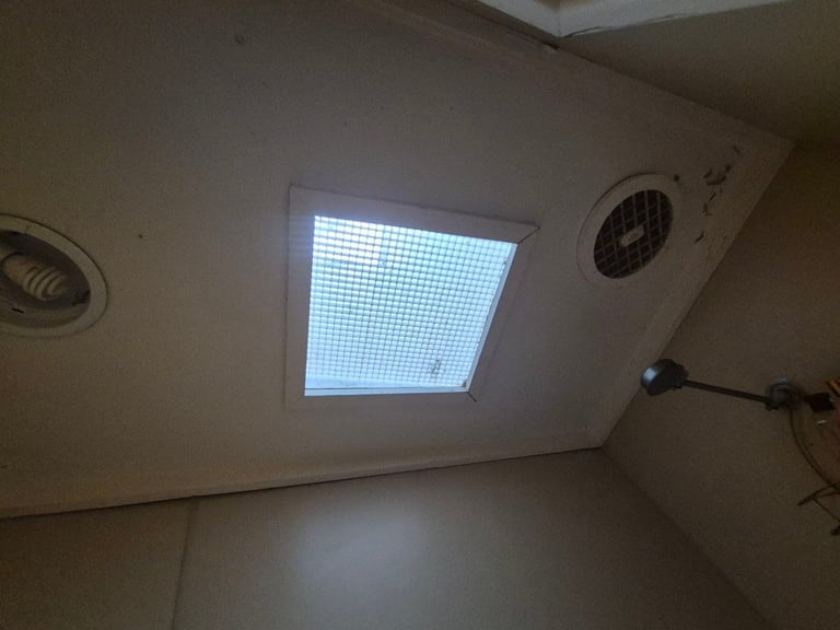 Interior ceiling view showing a bright skylight window with grid pattern, round vents, and electrical outlet on beige walls