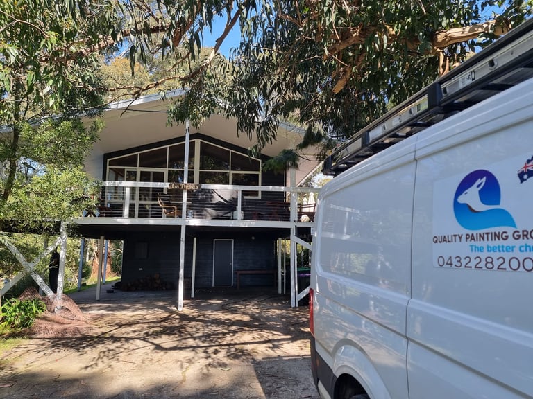 A white van from a painting company parked outside a two-story house with a carport, surrounded by trees and overgrown vegetation.