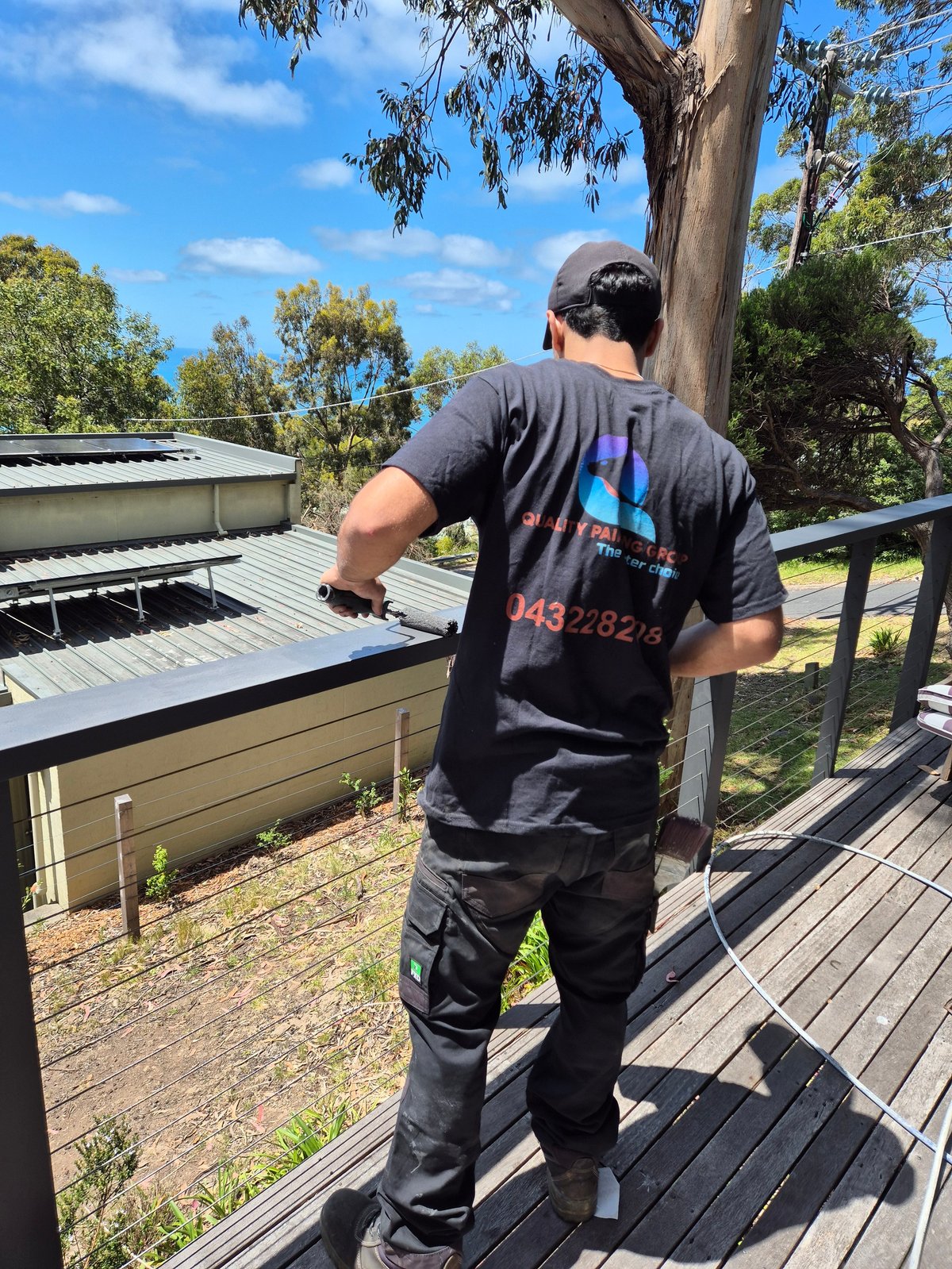 Person in black shirt and cap standing on wooden deck overlooking rural property with metal roofed structures and eucalyptus trees under blue sky
