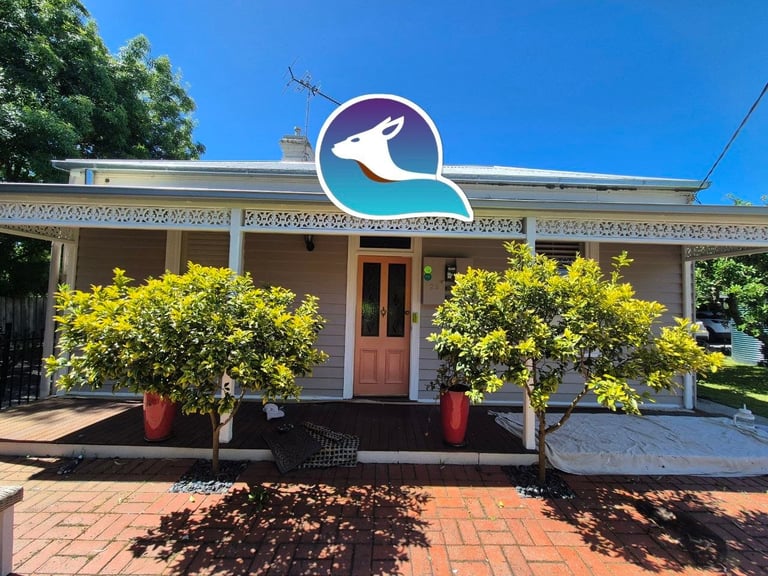 Storefront with beige facade, brown door, decorative trim, and two yellow flowering shrubs in red pots. Large blue and white logo above entrance against clear sky.