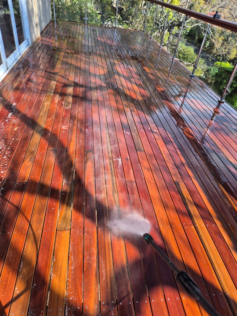 Person power washing a red-orange wooden deck, with water spray visible and green trees in the background