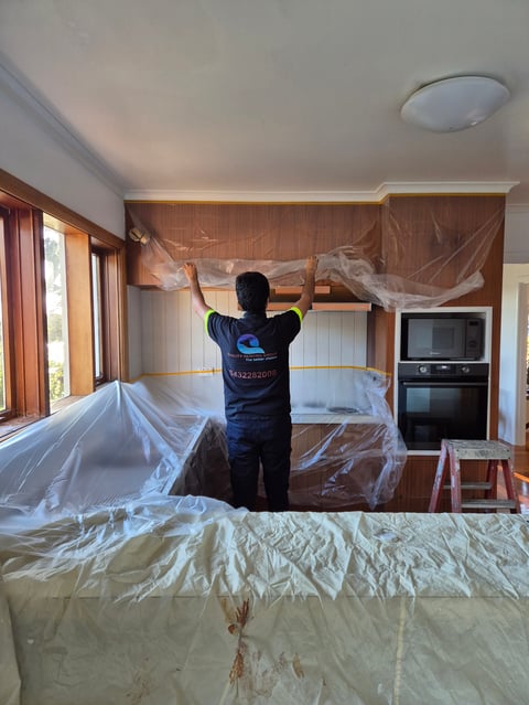 Worker in protective suit covering kitchen cabinetry and appliances with plastic sheeting during renovation