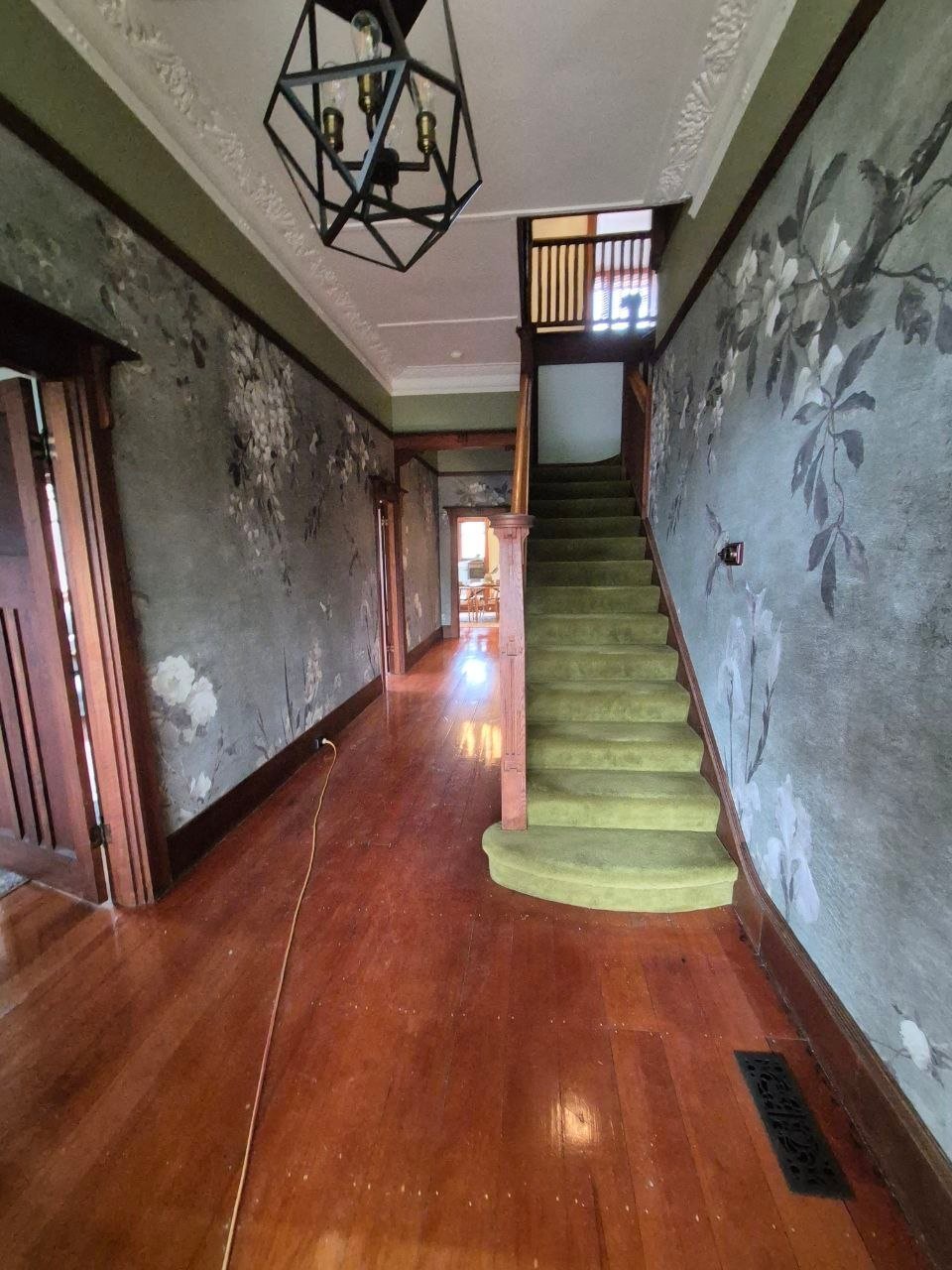 Narrow hallway with deteriorating green walls, wooden floor, geometric ceiling light, and curved green staircase
