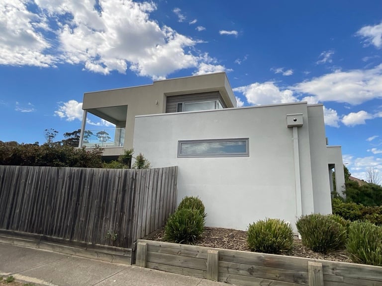 Modern white cubic house with metal fence, landscaped front garden, and blue sky with clouds