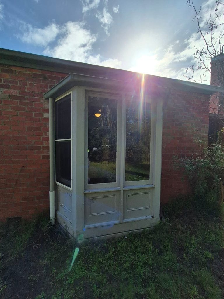A white-framed glass door and window structure attached to a red brick building, photographed in bright sunlight with sky and clouds visible above