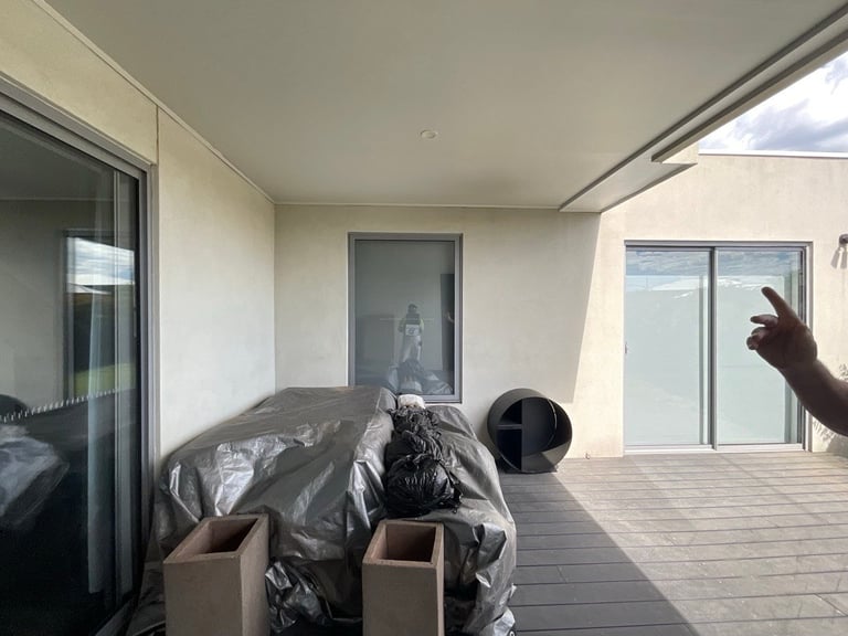 Modern balcony or terrace with white walls, large windows, gray composite flooring, and storage items covered with black plastic