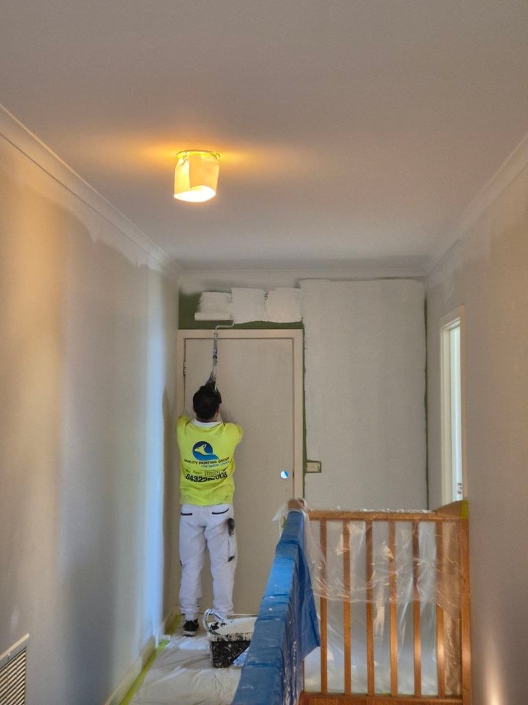 Person in yellow shirt standing on ladder reaching toward ceiling light fixture in white hallway with wooden railing