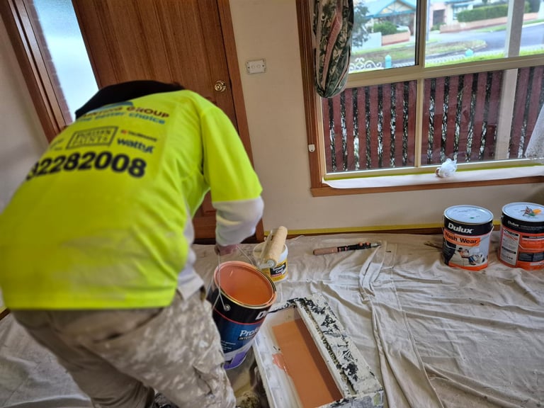 Worker in neon yellow hi-vis shirt preparing painting supplies and equipment on drop cloth indoors with window view.
